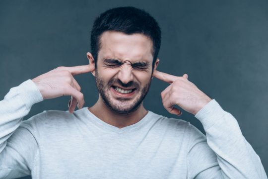 Too loud sound! Frustrated young man expressing negativity while covering ears with hands and standing against grey background Too loud sound! Frustrated young man expressing negativity while covering ears with hands and standing against grey background